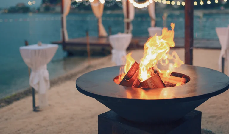 Fire pit with vibrant flames on a sandy shore, surrounded by covered tables, near Lake Norman townhomes at dusk.
